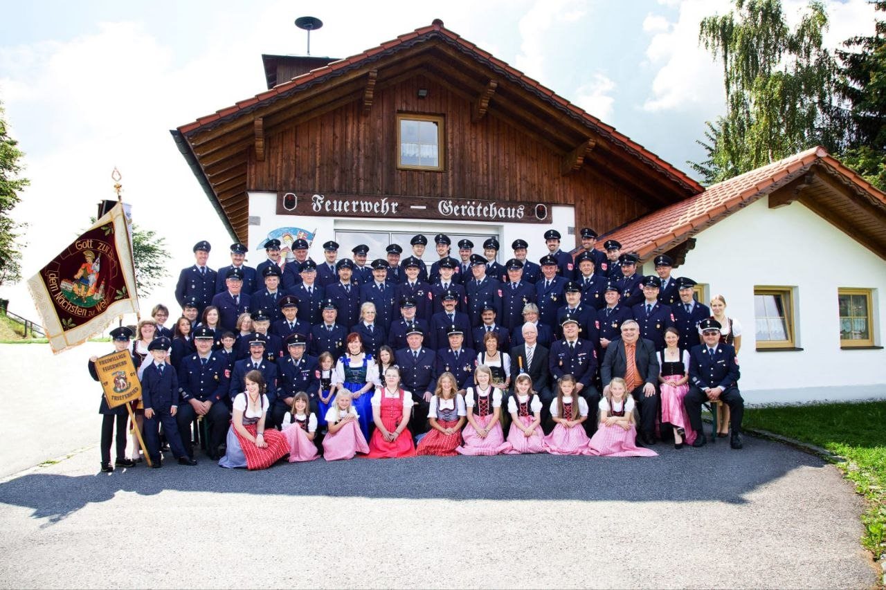 Gruppenbild der Freiwilligen Feuerwehr Triefenried in Uniform mit Vereinsfahne vor dem Feuerwehrhaus